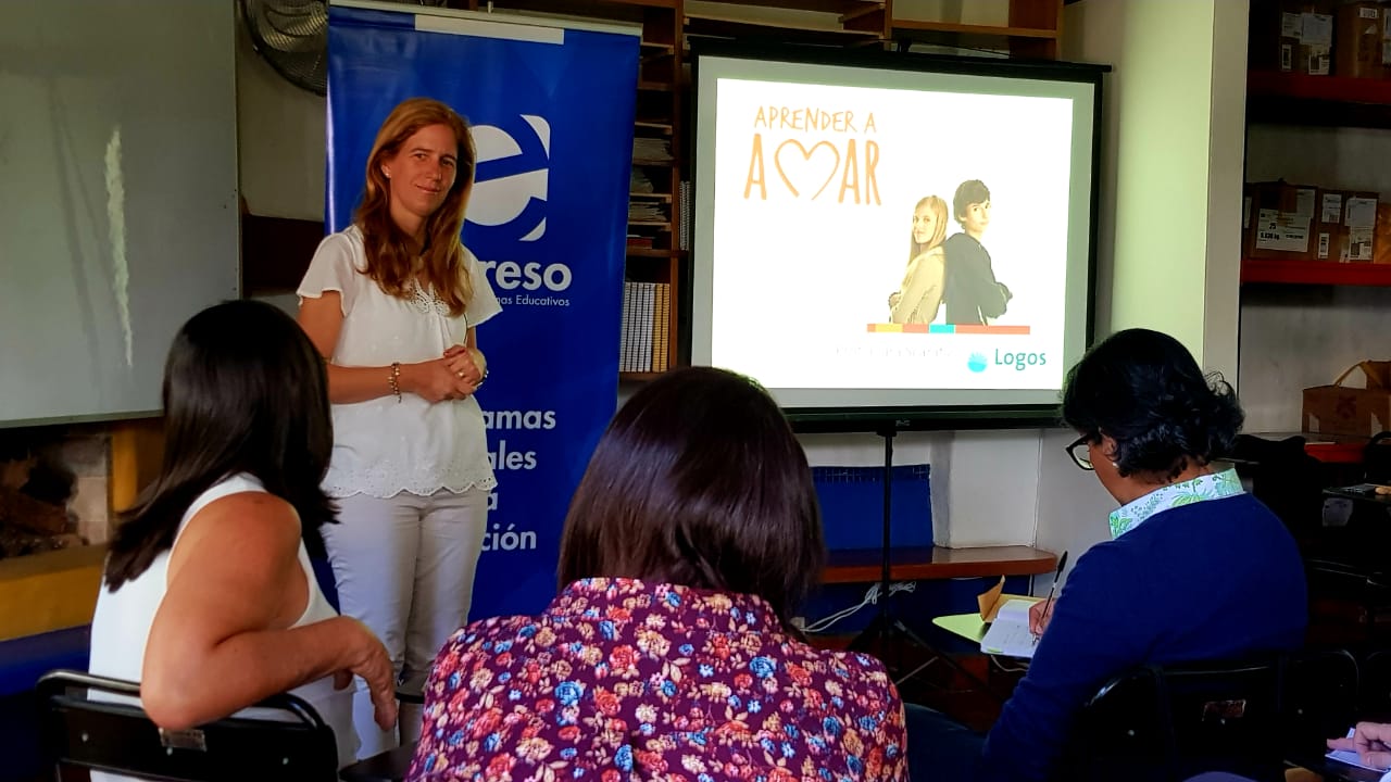 Una ponente frente a un grupo de personas durante una presentación del programa Aprender a Amar con una diapositiva proyectada al fondo Una ponente frente a un grupo de personas durante una presentación del programa Aprender a Amar con una diapositiva proyectada al fondo