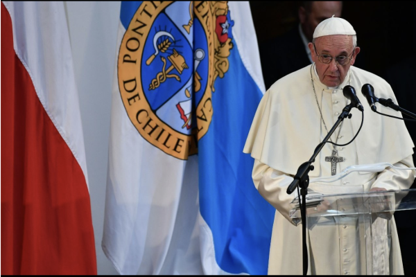 El Papa Francisco pronunciando un discurso tras un atril, con la bandera de Chile y el estandarte de la Pontificia Universidad Católica de Chile de fondo