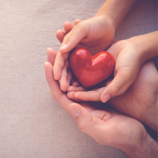 Primer plano de las manos de un adulto y un niño sosteniendo juntos un corazón rojo de madera, simbolizando el cuidado de la vida y el amor familiar.